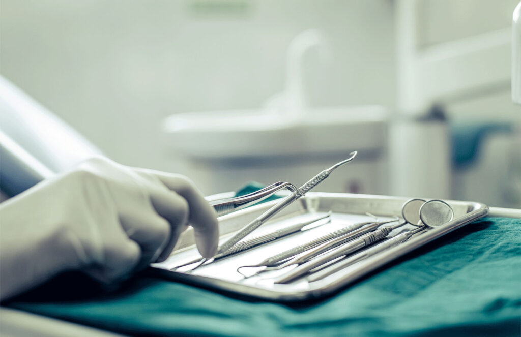 A gloved hand reaches toward a metal tray holding neatly arranged dental instruments in a clinical setting. The background shows a clean dental workspace with equipment softly out of focus.