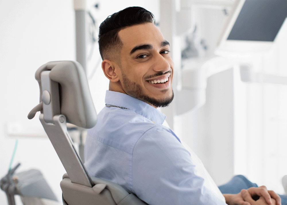 A person sits comfortably in a dental chair wearing a light blue shirt, ready for treatment. Behind them, sleek tools and a monitor hint at a clean, modern clinical setup.