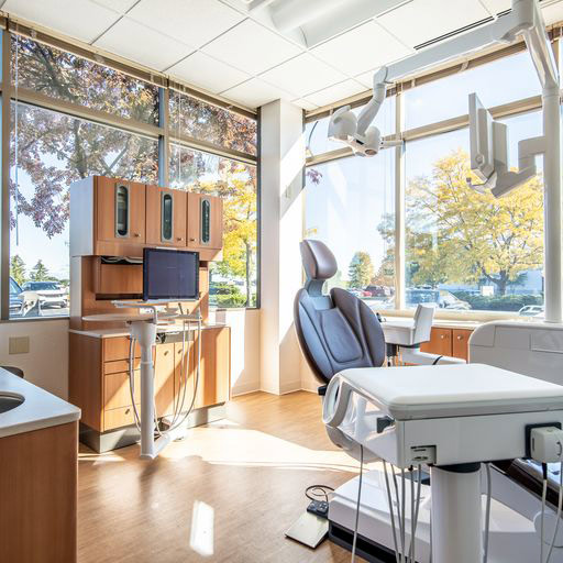 Dental exam room at Glo Dental Studio in Waukesha, WI. A clean, modern dental office features a patient chair surrounded by tools and equipment. Sunlight pours in through large windows with a view of trees in autumn and parked cars outside.