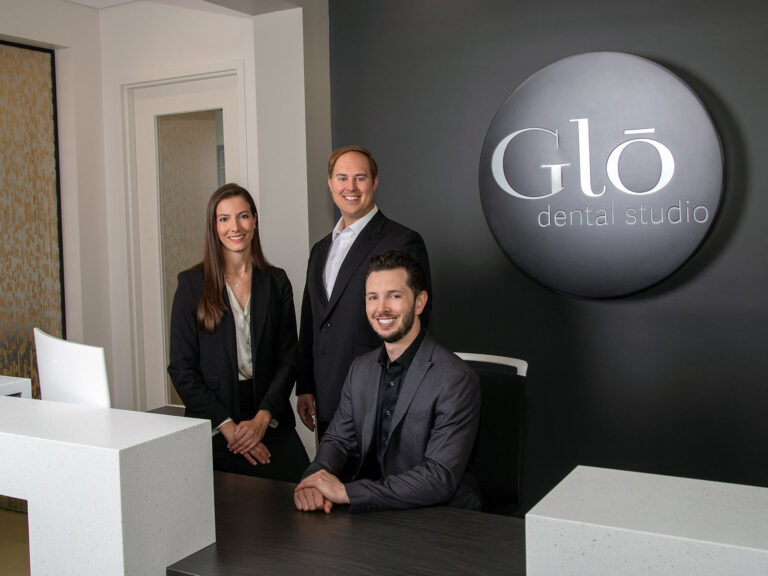 Dr. Emmerich, Dr. Jacoment, and Dr. Dwan of Glo Dental Studio in Waukesha, WI. Three team members at Glō Dental Studio pose confidently in the reception area, dressed in crisp business attire. A circular wall sign behind them reinforces the clinic's polished and professional brand.