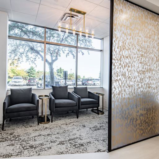 A modern waiting area features three dark gray armchairs, each paired with a small round side table with a gold base. A large window, abstract gold-and-white partition, and multi-bulb ceiling fixture add a sleek, upscale atmosphere to the space.