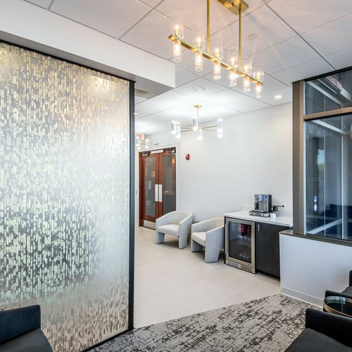 Image of the coffee station at Glo Dental Studio in Waukesha, WI. A stylish waiting area features two white armchairs beside a decorative wall and glass partition with abstract leaf-like patterns. Nearby, a coffee station and cylindrical chandelier enhance the clean, welcoming, and upscale ambiance.