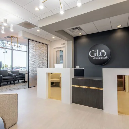 The reception area of Glō Dental Studio features a sleek white and dark wood desk accented by gold details, set against a black wall with the clinic’s logo. To the left, a cozy seating area and large window provide natural light, creating a bright, modern atmosphere.